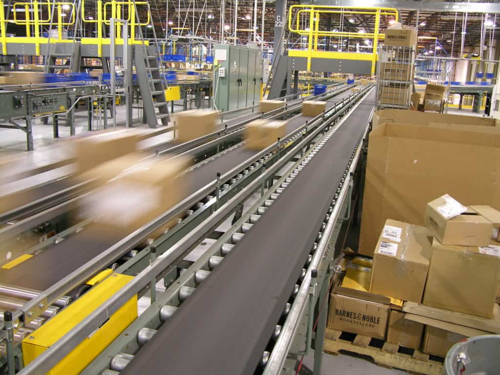 Vertical Lift Modules: How a VLM Works and Where It Fits Next to Conveyor 2 Cartons riding a belt conveyor past a pallet of staged cases in the Barnes & Noble distribution center integrated by Century Conveyor