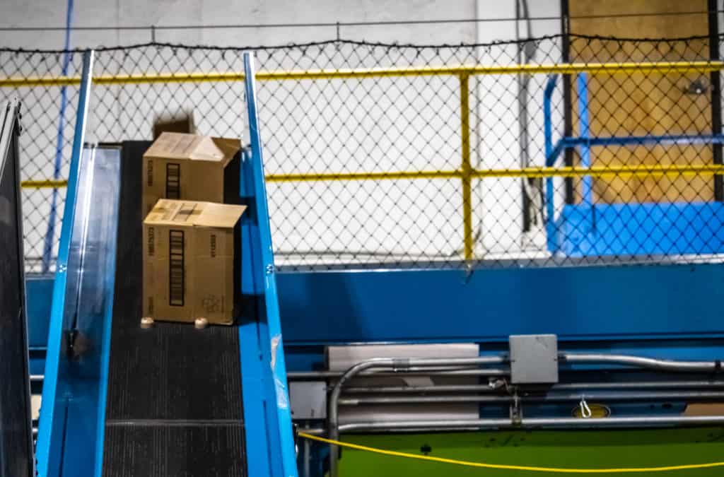 Conveyor Safety: Guarding, E-Stops, Pull Cords, and Lockout/Tagout 3 Conveyor safety fencing: a carton riding a blue incline belt conveyor with chain-link fencing behind the line and a yellow line running along the conveyor side in a co-packing facility installed by Century Conveyor