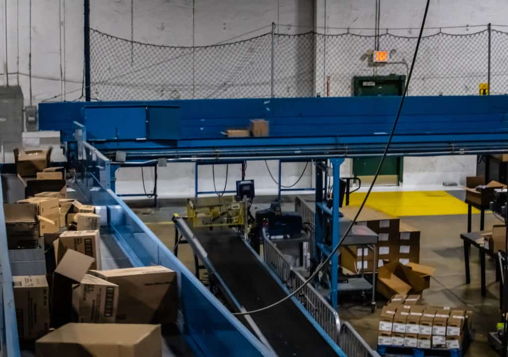 Conveyor Belt Tracking: Why Belts Wander and How Technicians Correct It 3 Blue-framed belt conveyors carrying empty cartons past pack stations in the co-packing and corrugate disposal facility, a line where conveyor belt tracking is checked often